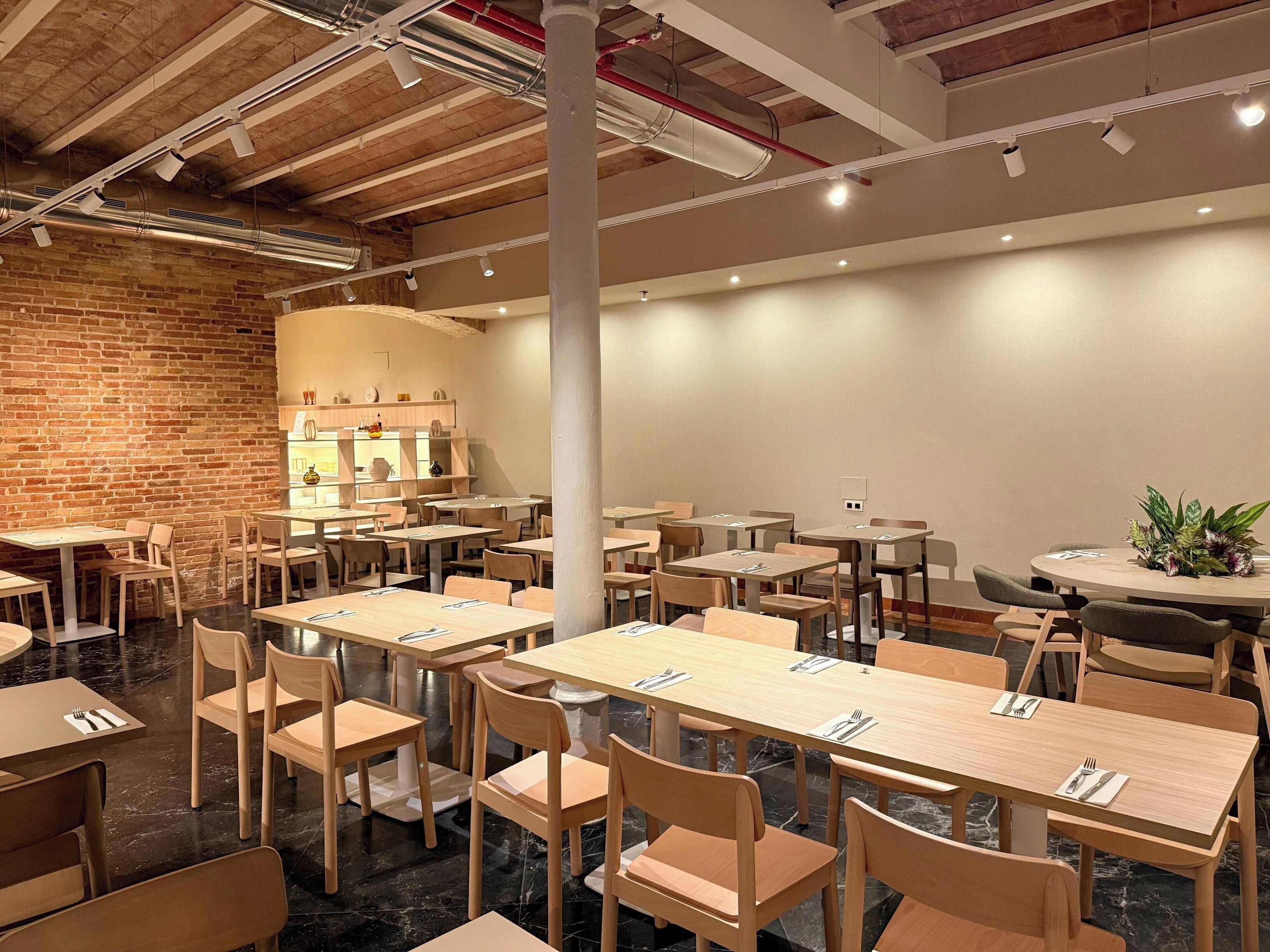 Un restaurante con paredes de ladrillo, mesas y sillas de madera, y un techo moderno con vigas y luminarias a la vista.