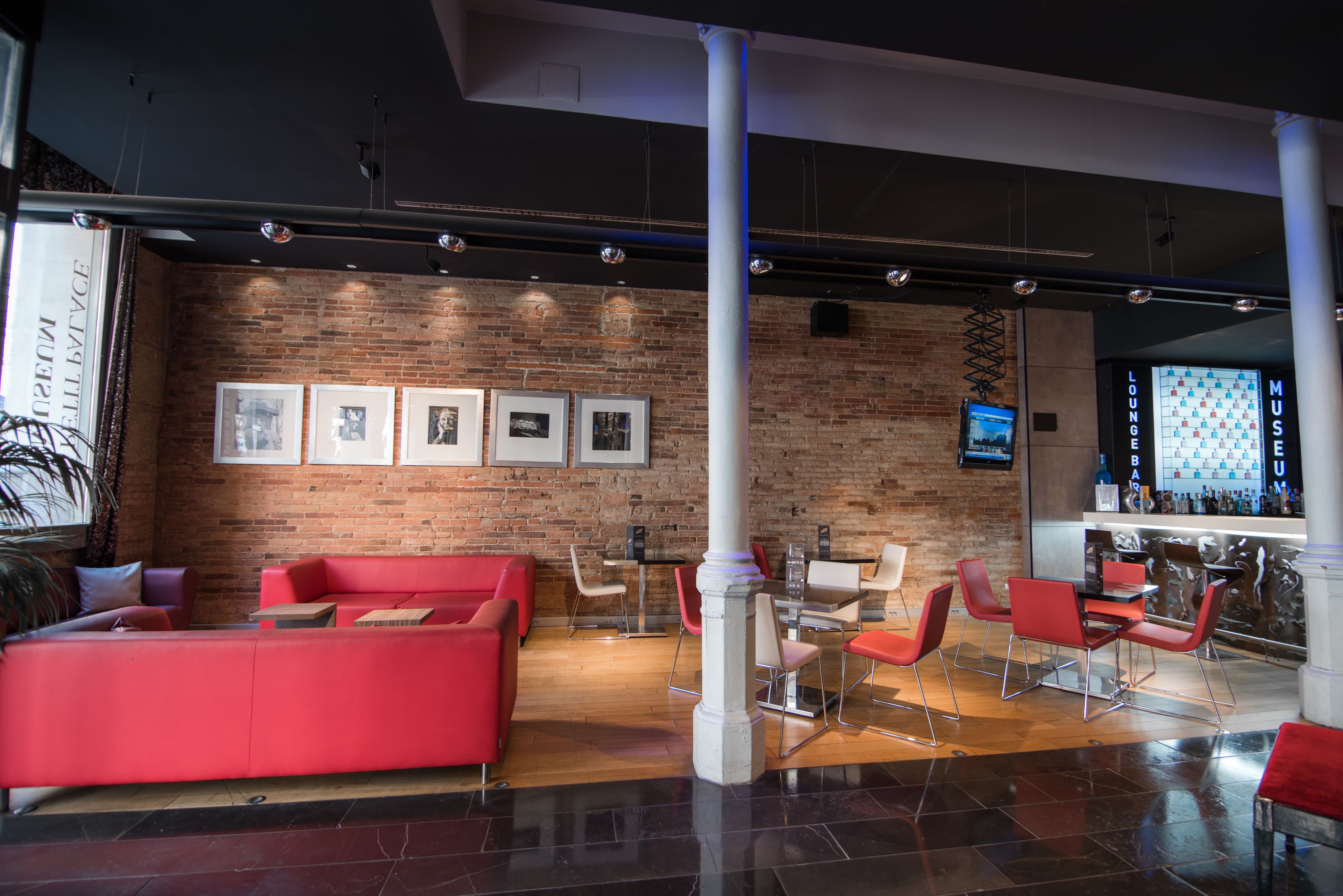 A modern lounge area with red seating, a brick wall with framed pictures, and a bar area with a screen displaying a game.