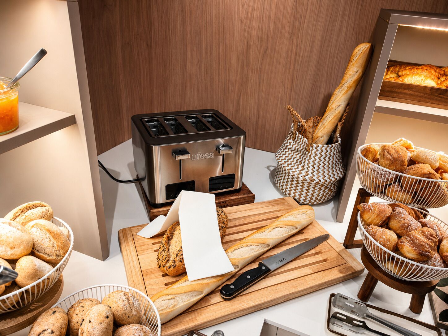 Una zona de cocina con un tostador, cestas de pan, una tabla de cortar con un cuchillo y una cesta de pasteles.