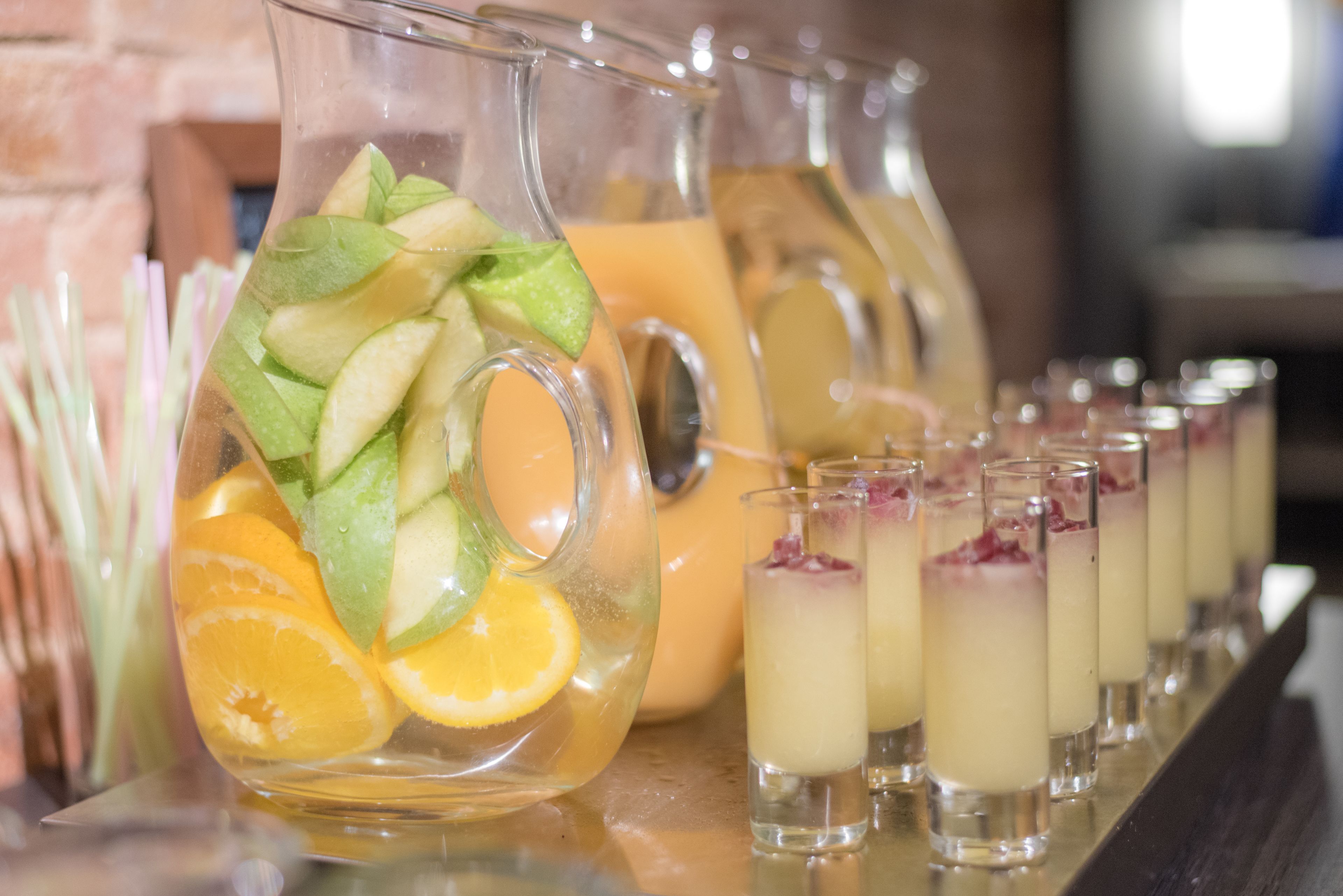 Una jarra llena de rodajas de fruta y una hilera de vasos pequeños con bebidas en capas.