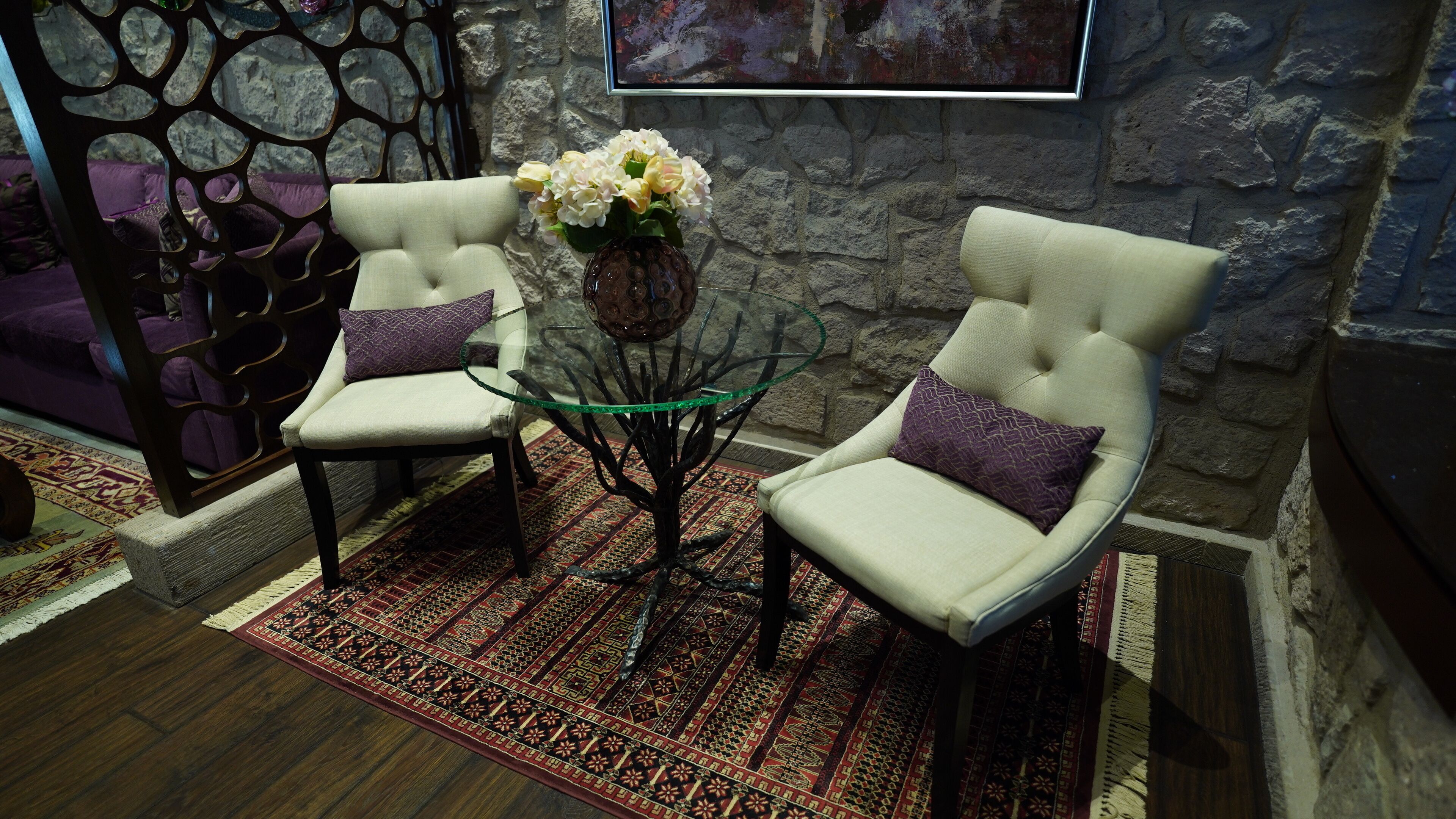 Una habitación con dos sillones blancos, una mesa de centro de vidrio, un jarrón con flores, una alfombra estampada y una pared de piedra con un cuadro.