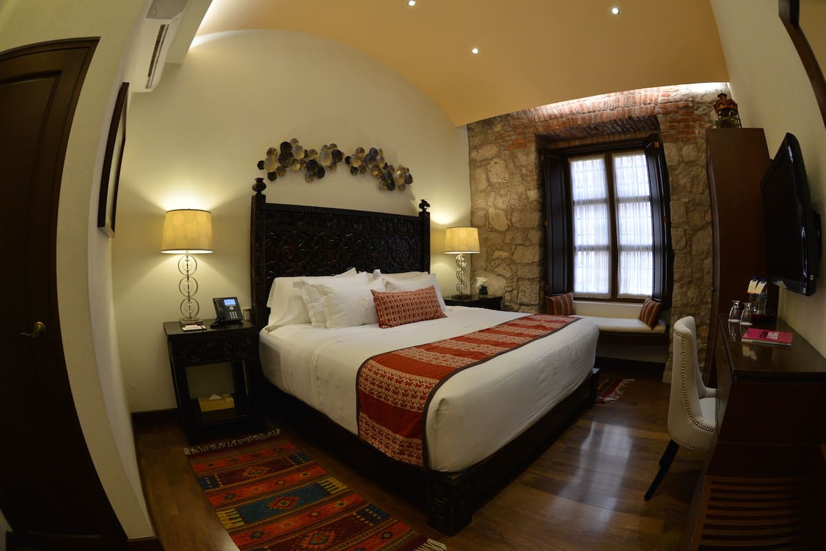 A bedroom with a large bed, two bedside tables with lamps, a stone accent wall, a window with curtains, and a wooden floor.