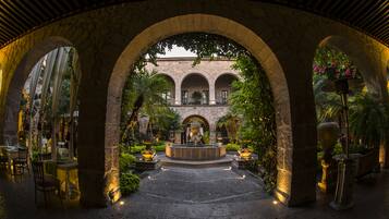 An outdoor courtyard with stone arches, a central fountain, and tables and chairs for dining.