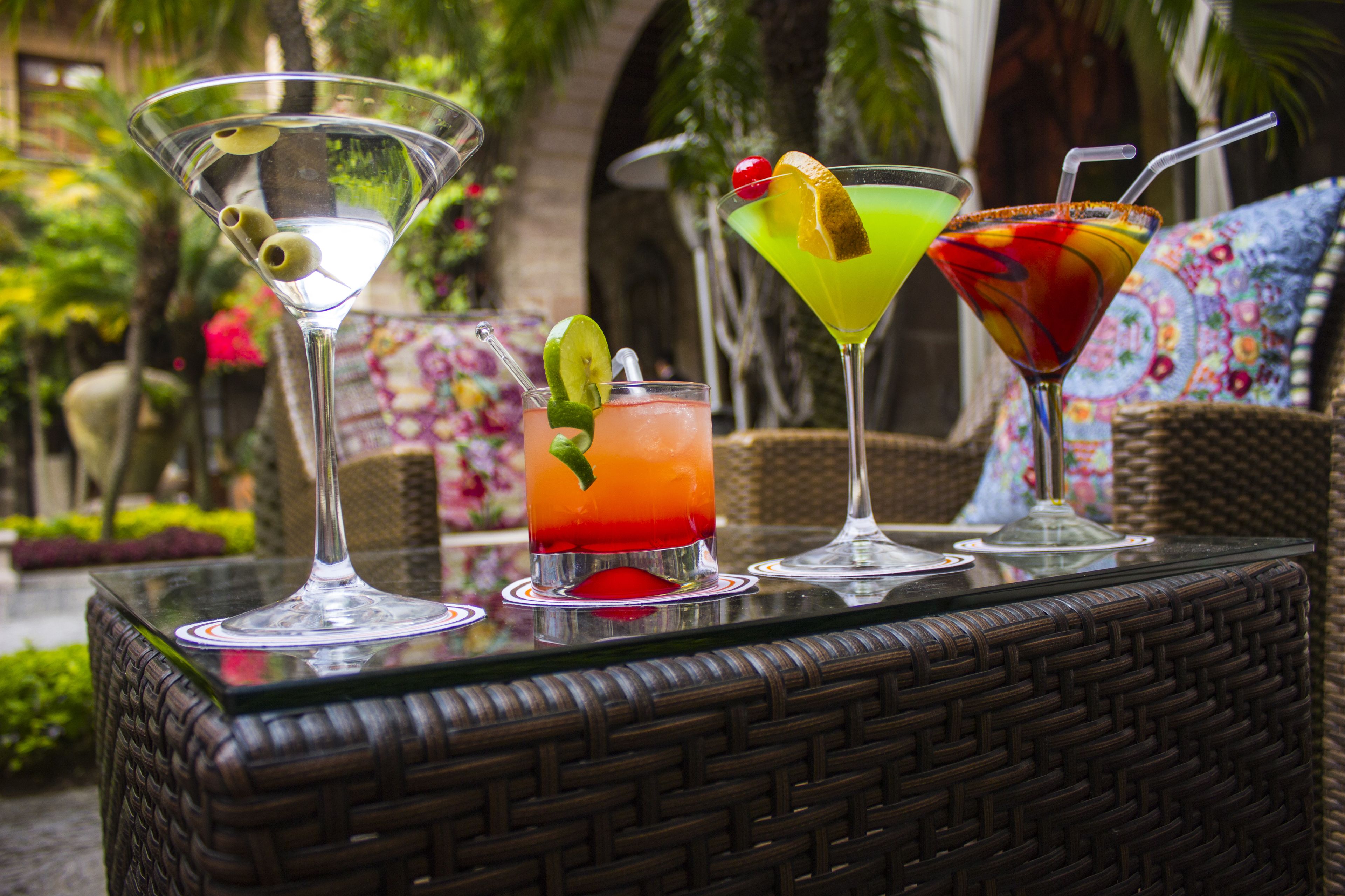 Una mesa con cuatro vasos de martini que contienen cócteles coloridos, sobre un fondo de vegetación exuberante y muebles de mimbre.