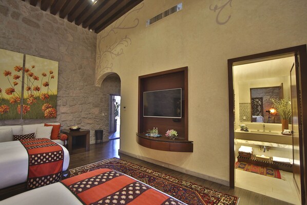 Habitación de hotel con una cama grande, un televisor de pantalla plana montado en la pared, un muro de piedra y vista al baño a través de una puerta de vidrio.