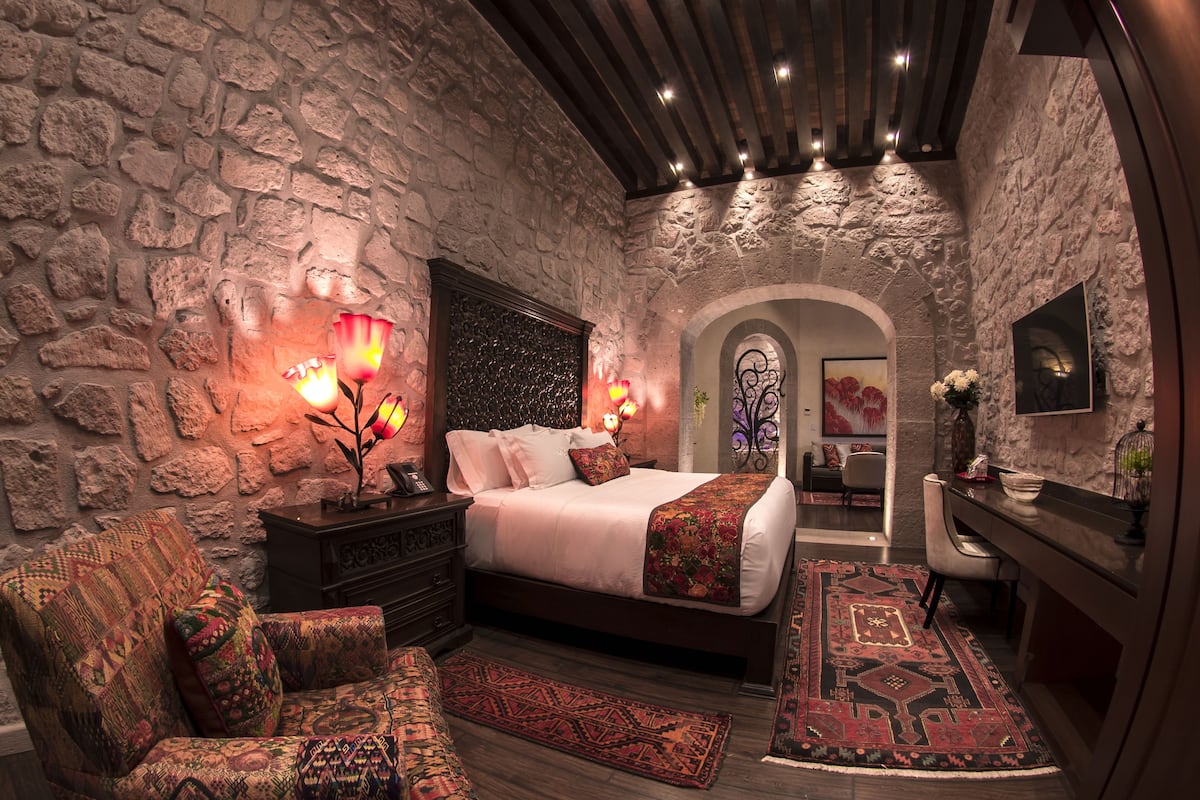 A bedroom with a stone wall, a bed with a floral bedspread, a nightstand with a lamp, a patterned armchair, and a red and black rug.