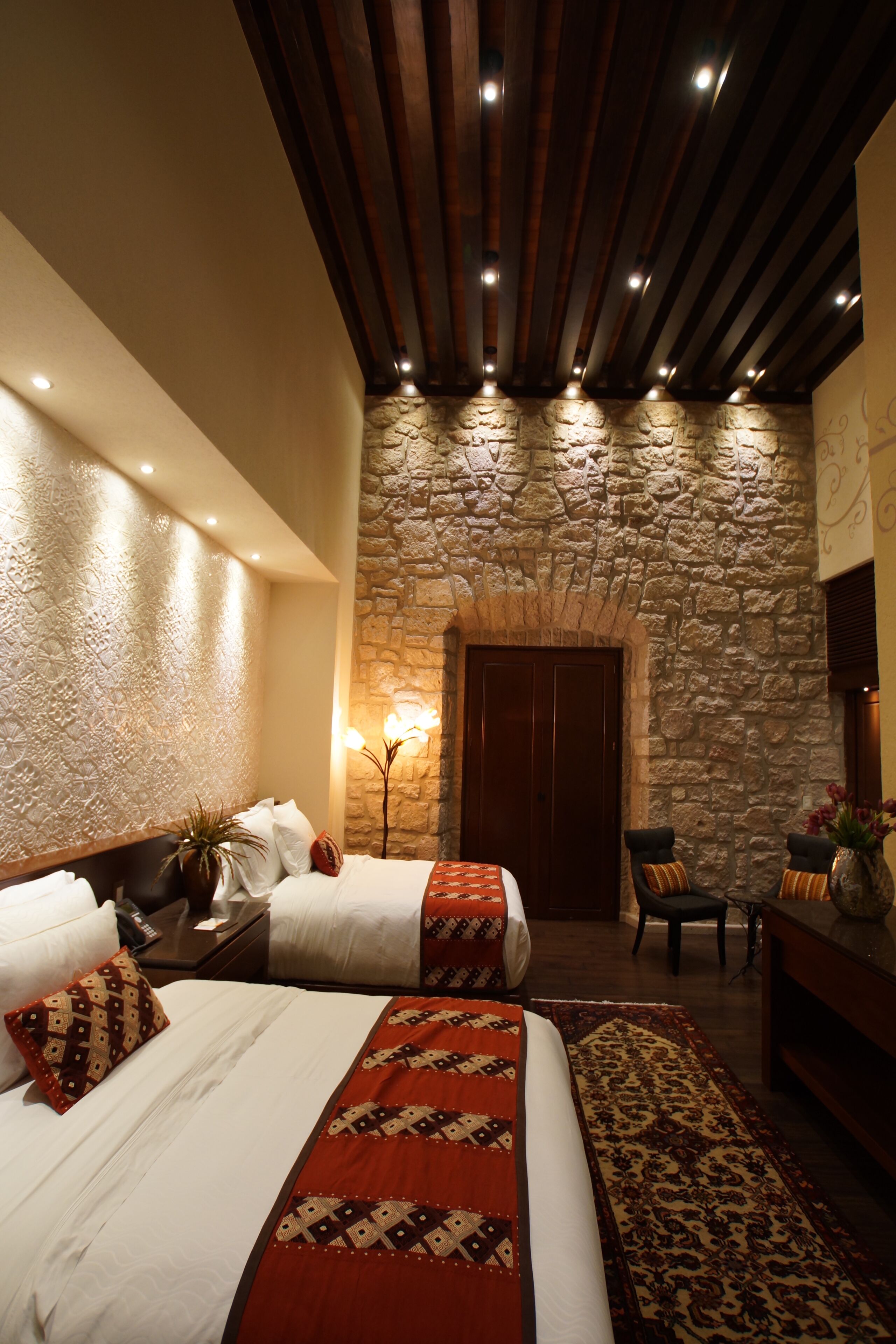Habitación de hotel con pared de piedra, dos camas con ropa de cama roja y blanca, techo de madera y un arco de piedra.