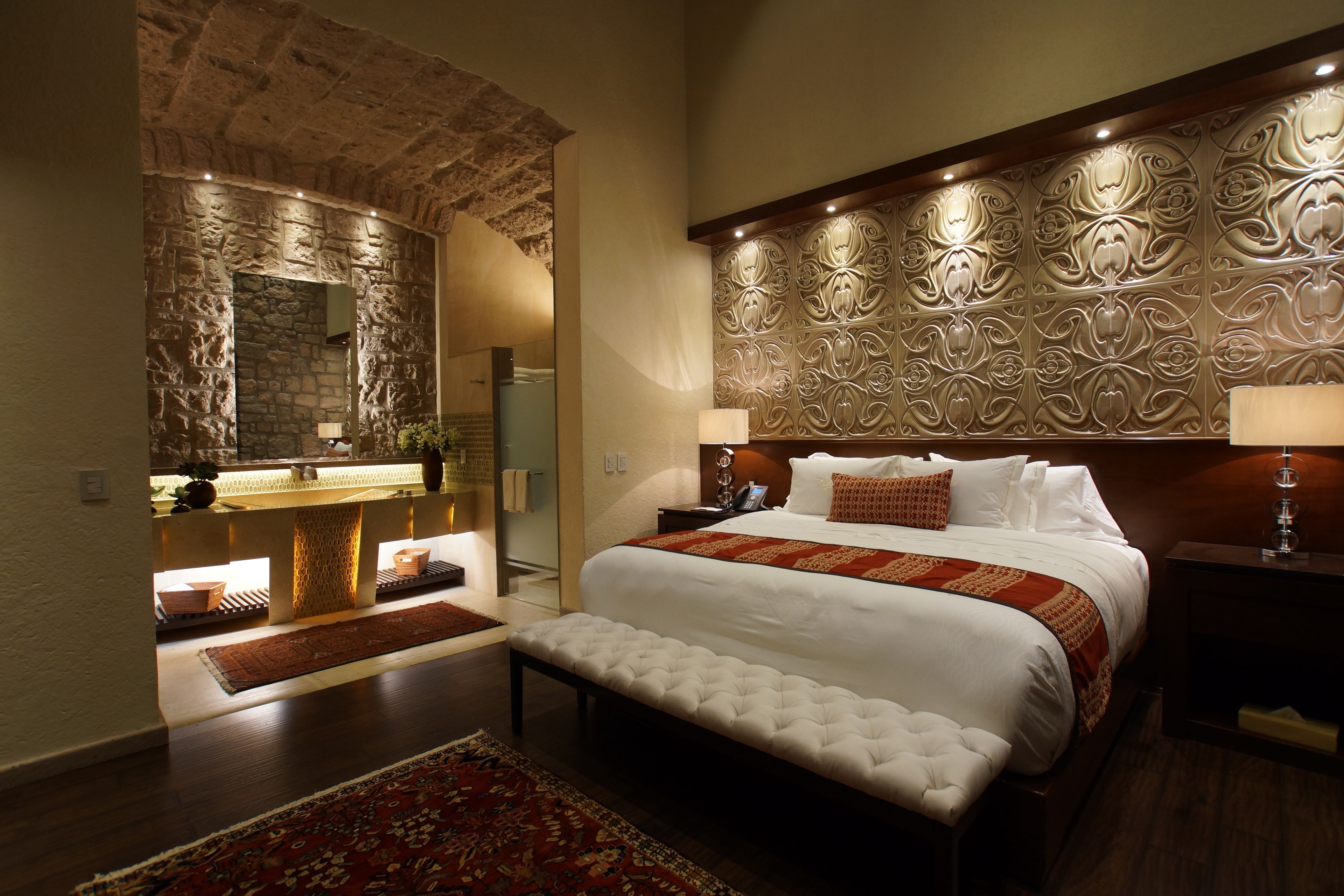 A bedroom with a stone wall, a large bed with a red and white bedspread, a bench, and a nightstand with a lamp.