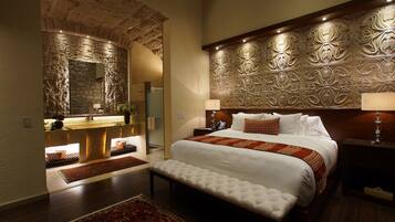 A bedroom with a stone wall, a large bed with a red and white bedspread, a bench, and a nightstand with a lamp.