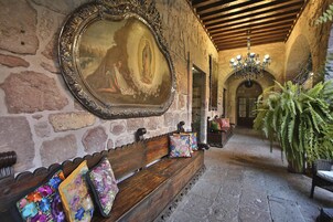 A stone-walled corridor with a wooden bench, a large ornate mirror, and a chandelier.