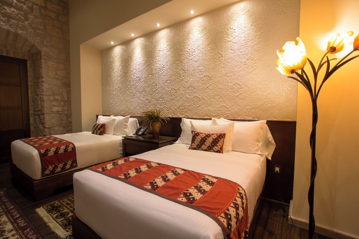 Habitación de hotel con dos camas, pared de piedra, techo con textura y un jarrón con flores.