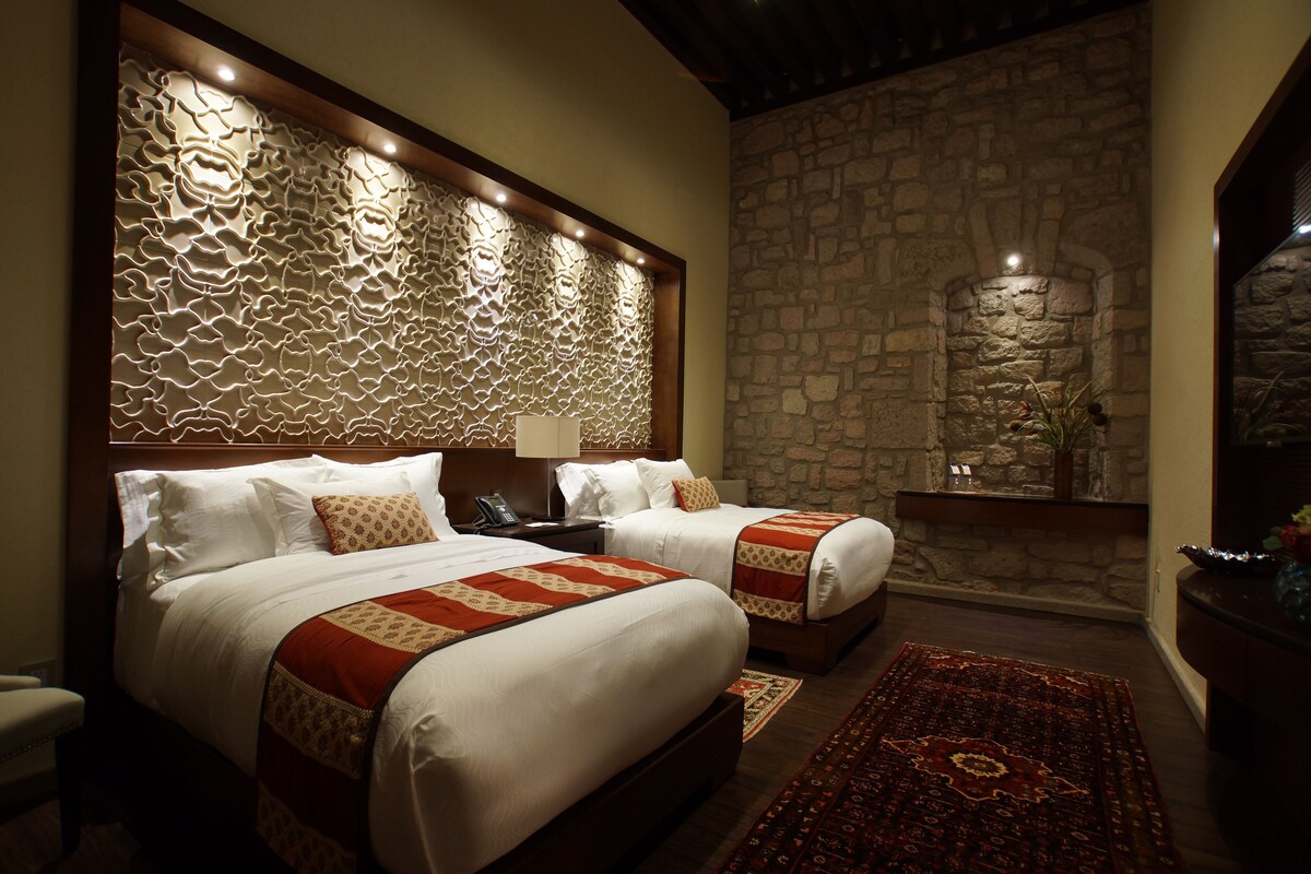 Habitación de hotel con dos camas, pared de piedra y cabecero decorativo.