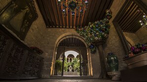 Un arco de entrada da a un patio con una fuente, rodeado de muros de piedra y un techo de madera con adornos de vidrio colgantes.