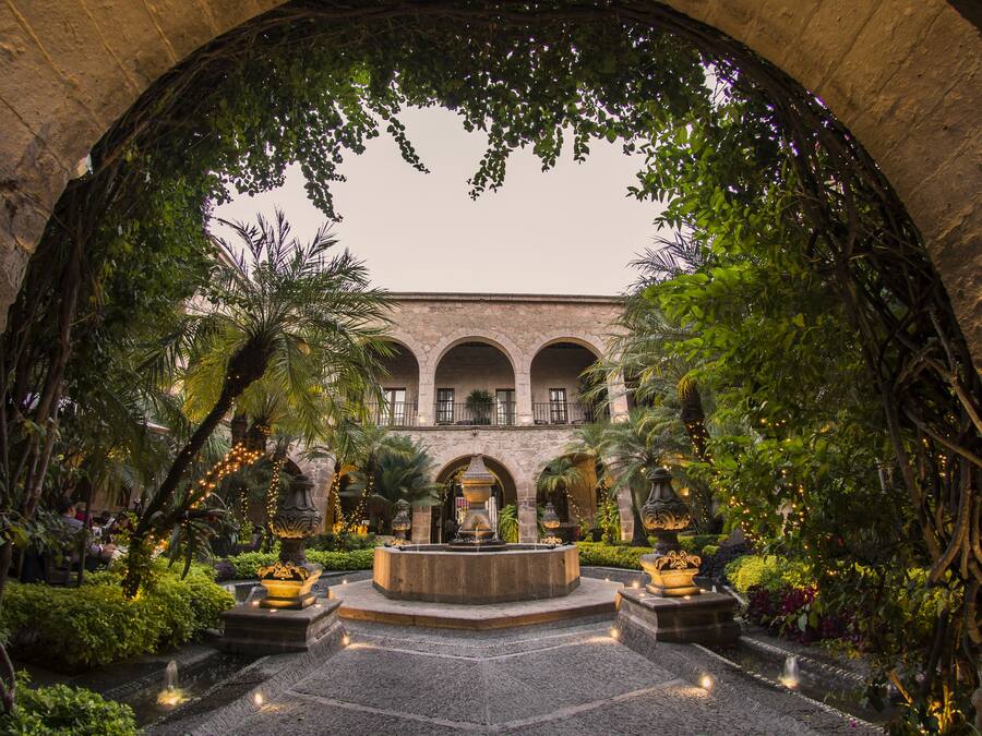Um pátio em arco com uma fonte central, rodeado por vegetação exuberante e arquitetura em pedra.