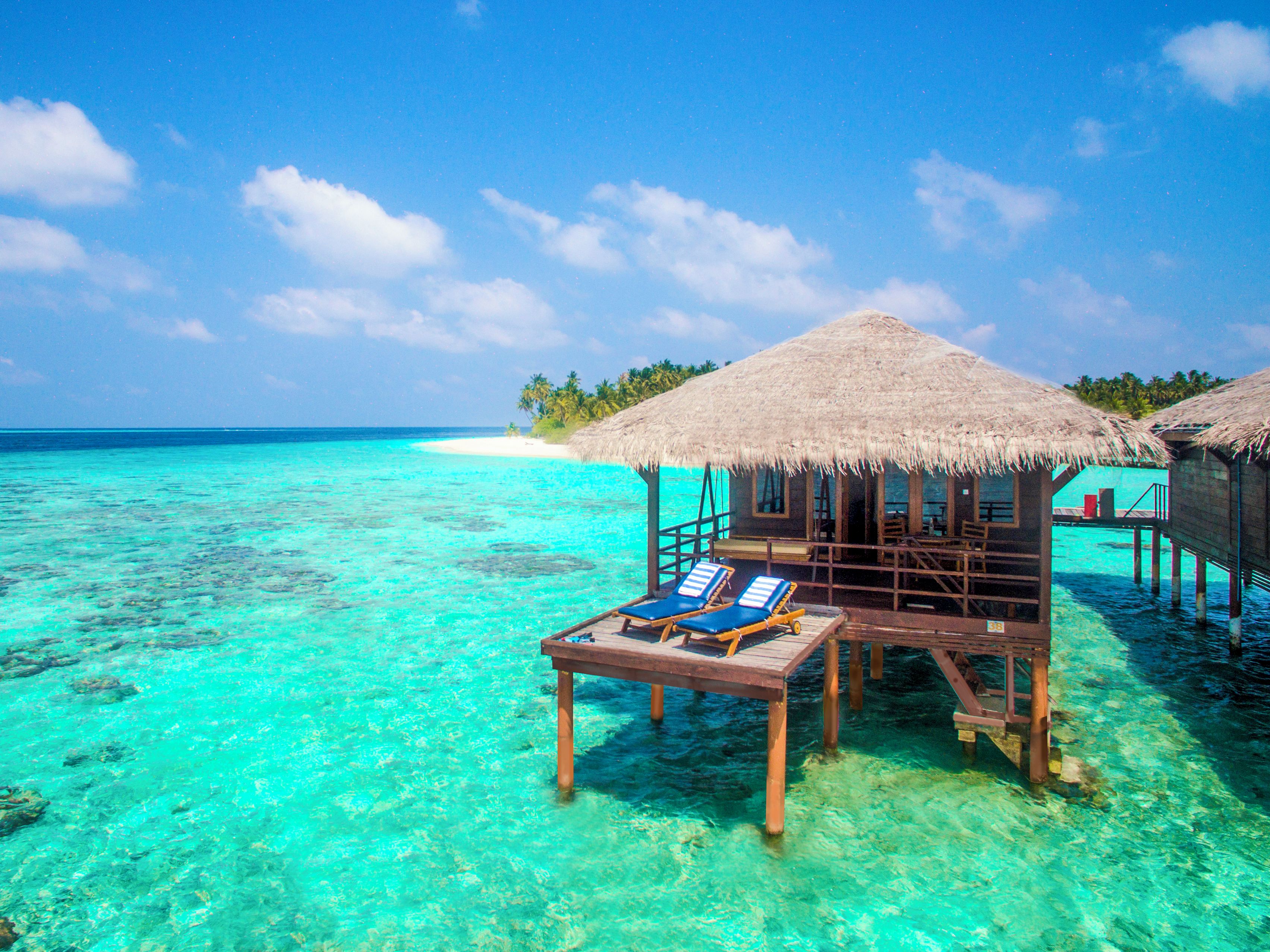 Overwater bungalows with thatched roofs and wooden floors, clear turquoise water, and a white sandy beach in the background.