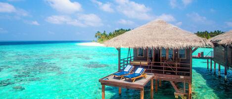 Overwater bungalows with thatched roofs and wooden floors, clear turquoise water, and a white sandy beach in the background.