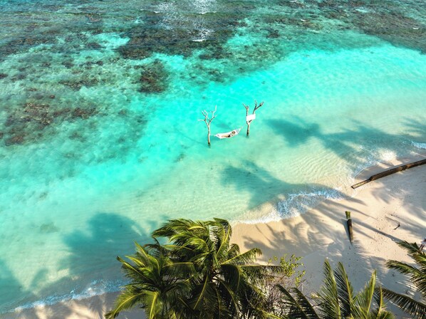 Vista aérea de una playa con agua turquesa cristalina, una hamaca atada entre dos árboles y palmeras en primer plano.