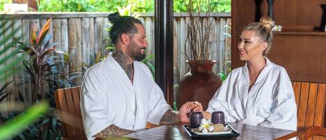 A couple in white robes seated at a wooden table with a tray of snacks and a flower.