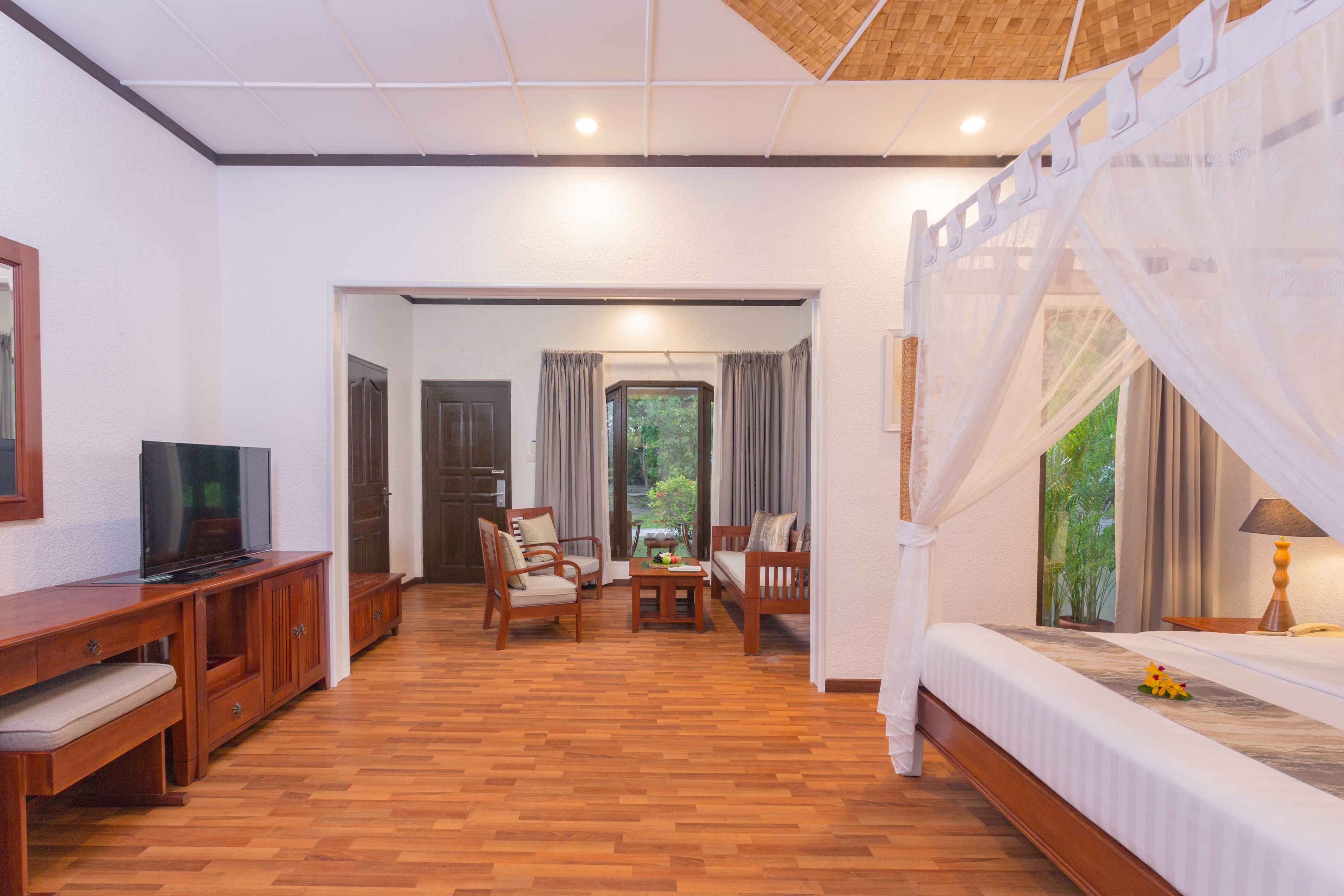 A hotel room with a wooden floor, a TV on a wooden cabinet, a bed with a canopy, and a view of greenery outside.