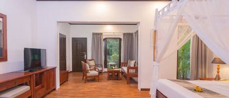A hotel room with a wooden floor, a TV on a wooden cabinet, a bed with a canopy, and a view of greenery outside.