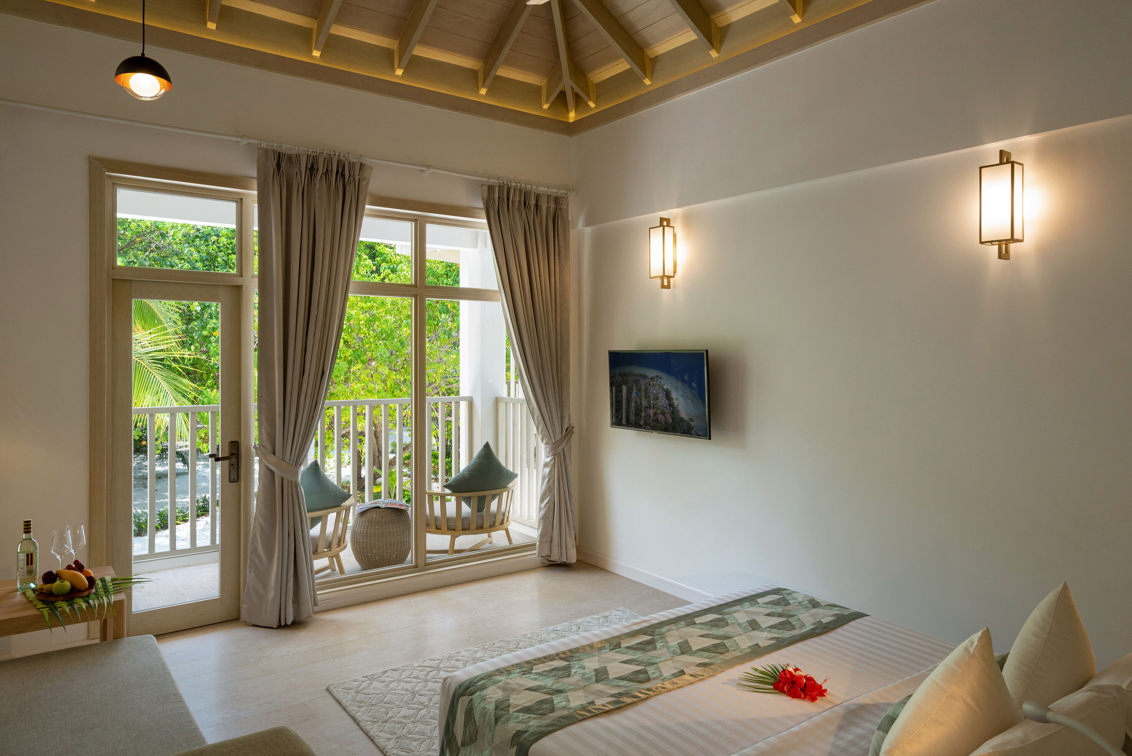 A bedroom with a bed, a window with curtains, a chair, a table with a bottle and fruit, and a painting on the wall.