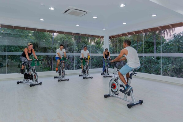 A group of people exercising on stationary bikes in a well-lit room with large windows and a view of greenery outside.