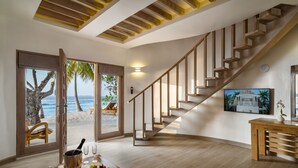 Un espacio amplio con una escalera de madera, un comedor con una mesa dispuesta para dos personas, y un gran ventanal con vistas a la playa y palmeras.