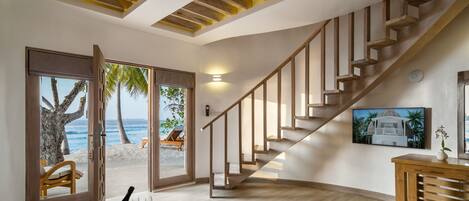 A spacious room with a wooden staircase, a dining area with a table set for two, and a large window offering a view of the beach and palm trees.