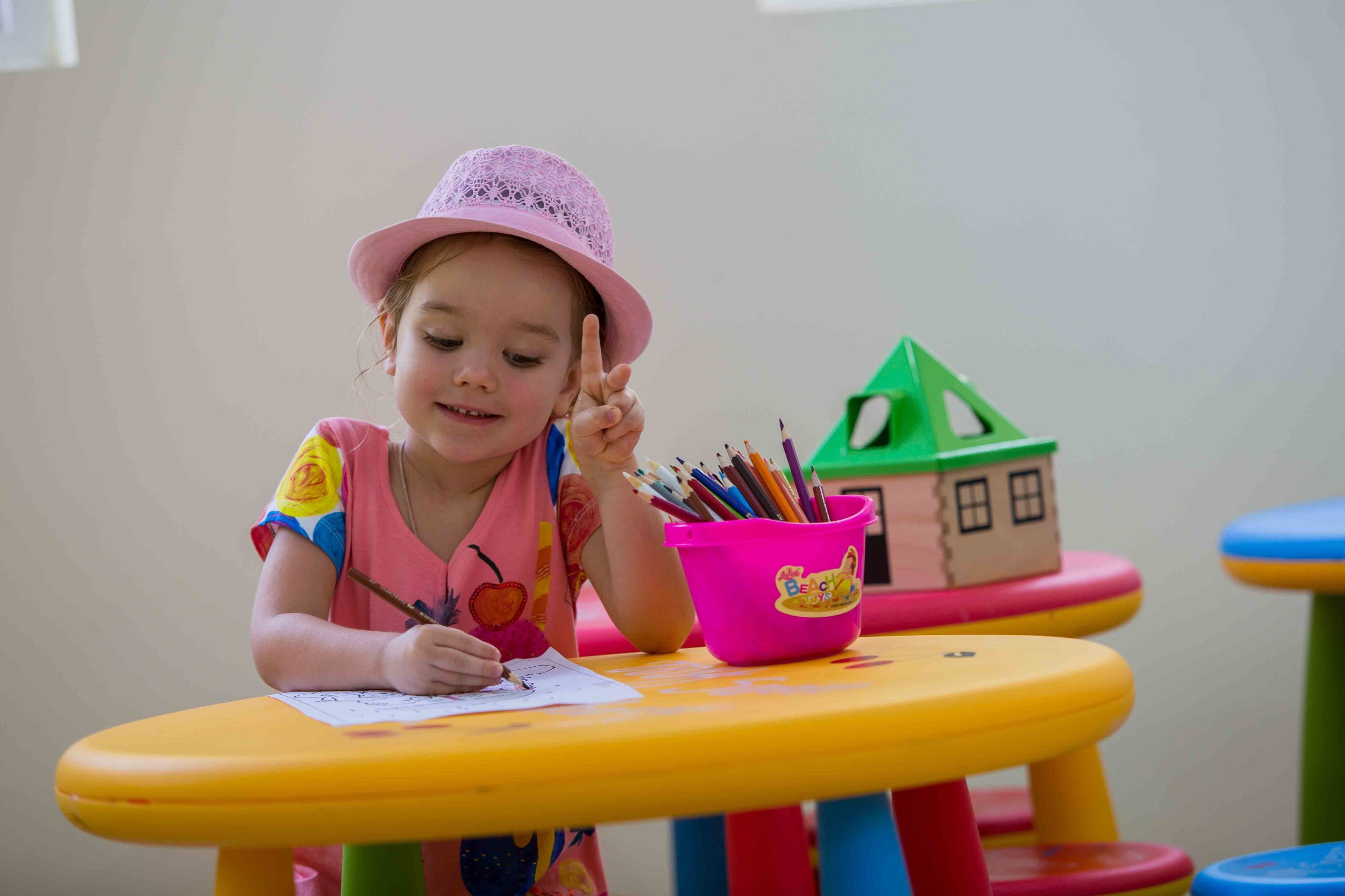 Un enfant est assis à une table colorée, portant un chapeau rose, et écrit à un bureau sur lequel se trouve un porte-crayons rose rempli de crayons de couleur.