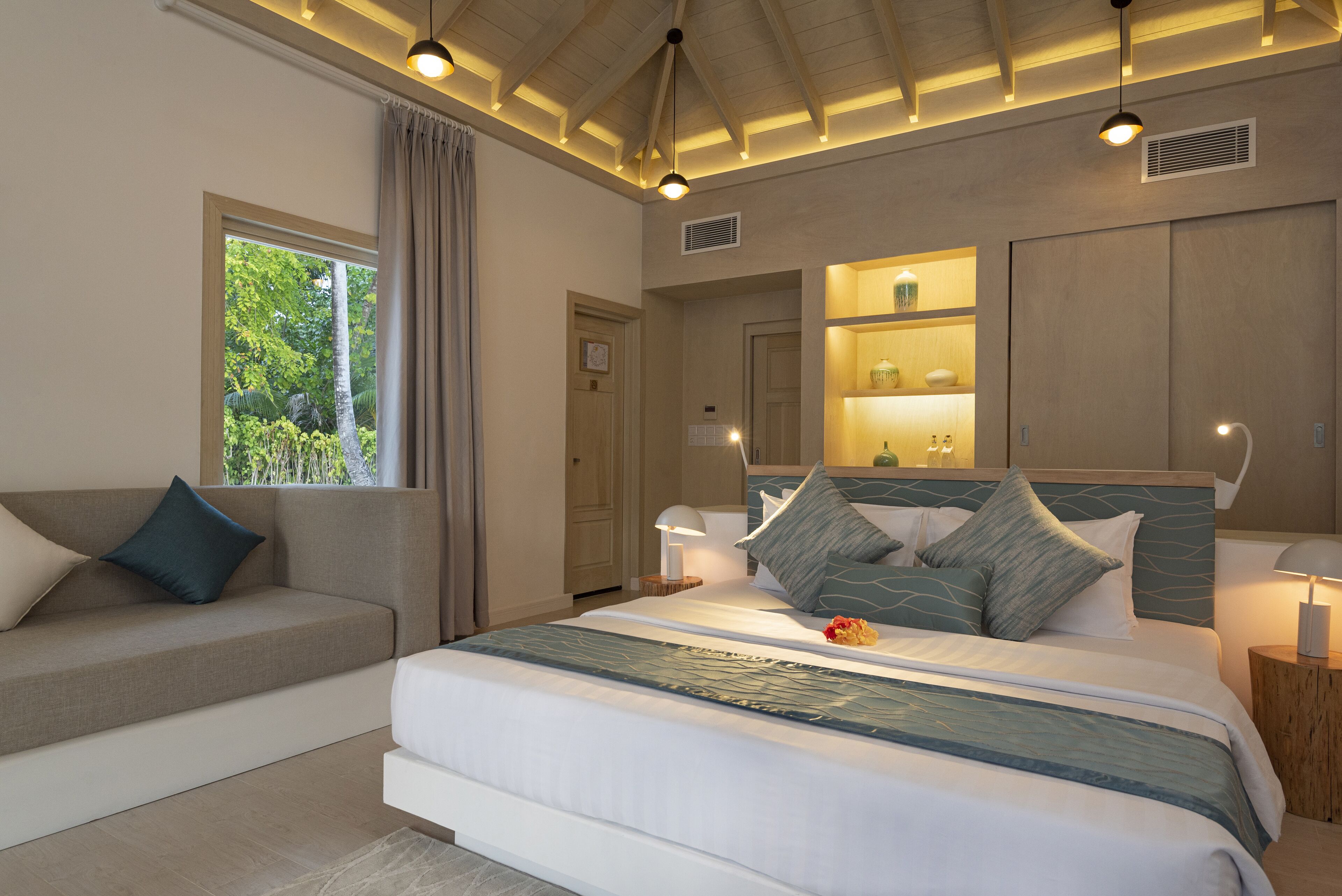 Un dormitorio moderno con una cama grande, un sofá, una ventana con cortinas y estanterías empotradas con objetos decorativos.