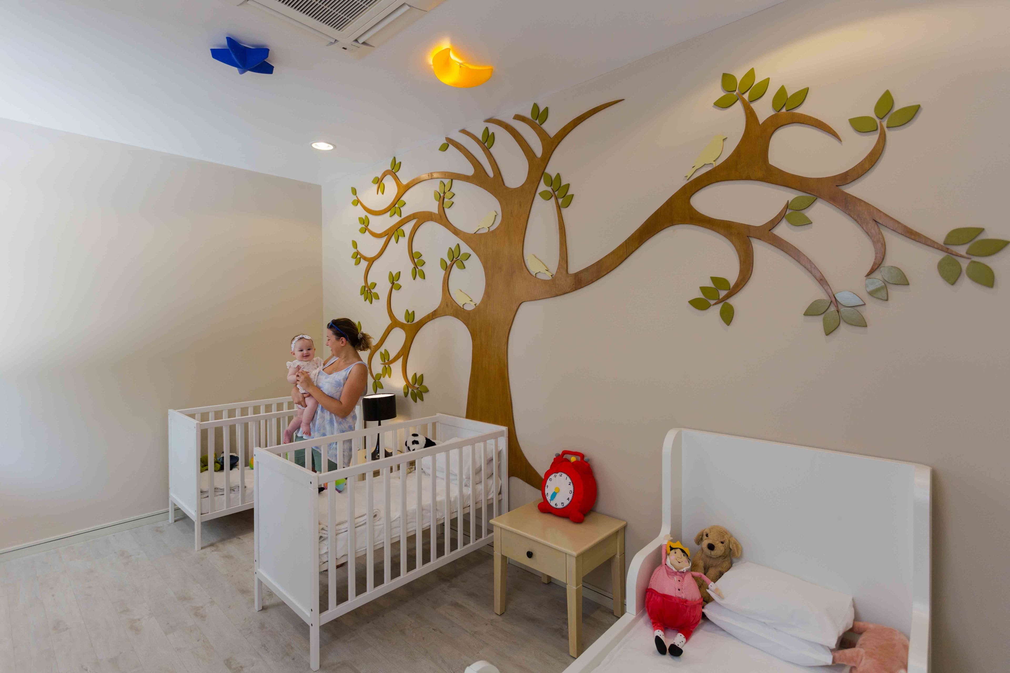 A baby room with two cribs, a small table, a wall mural of a tree, and a bed with a teddy bear.