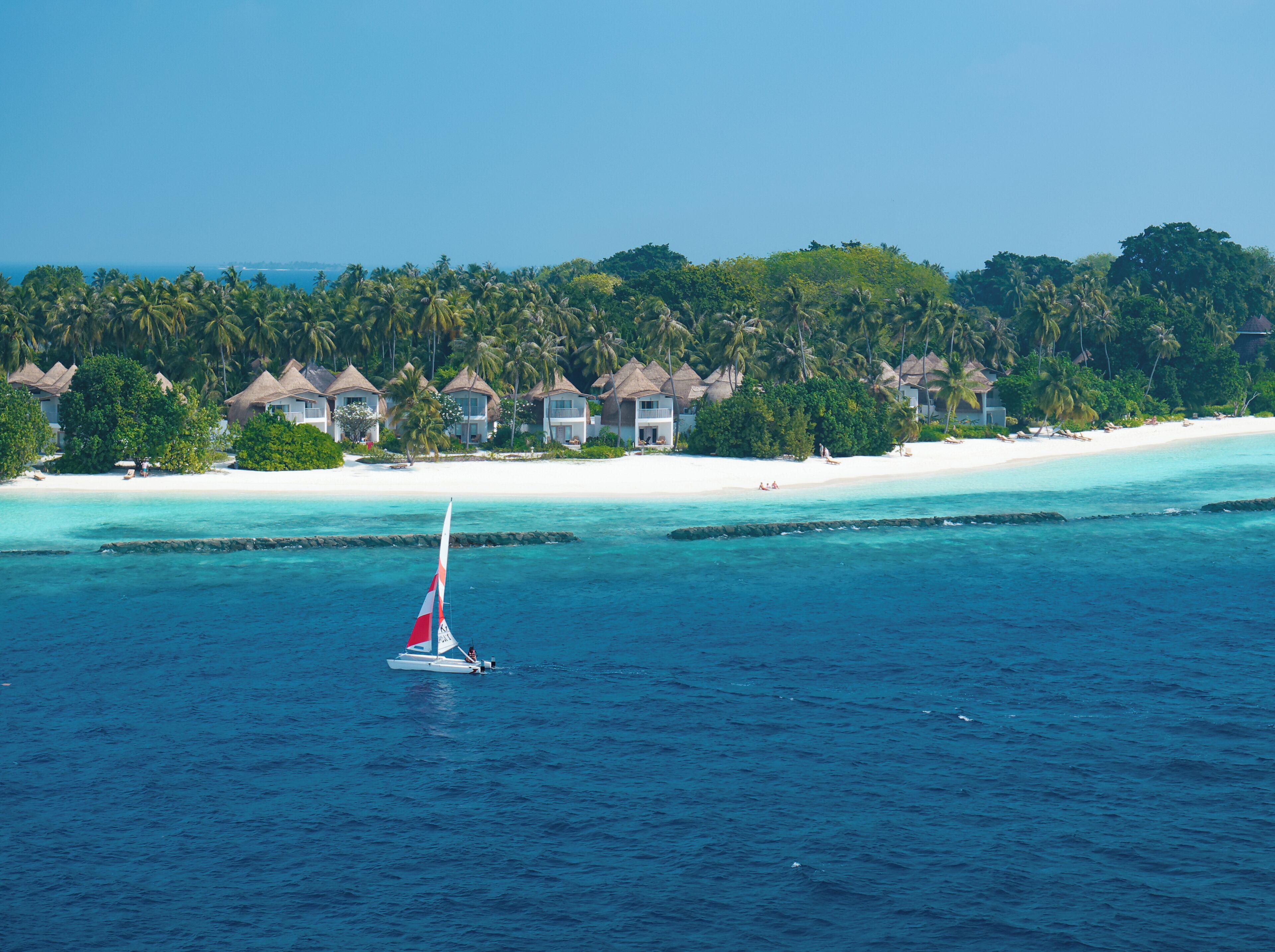 Een zeilboot op turquoise water, met een strand en overwater bungalows op de achtergrond.