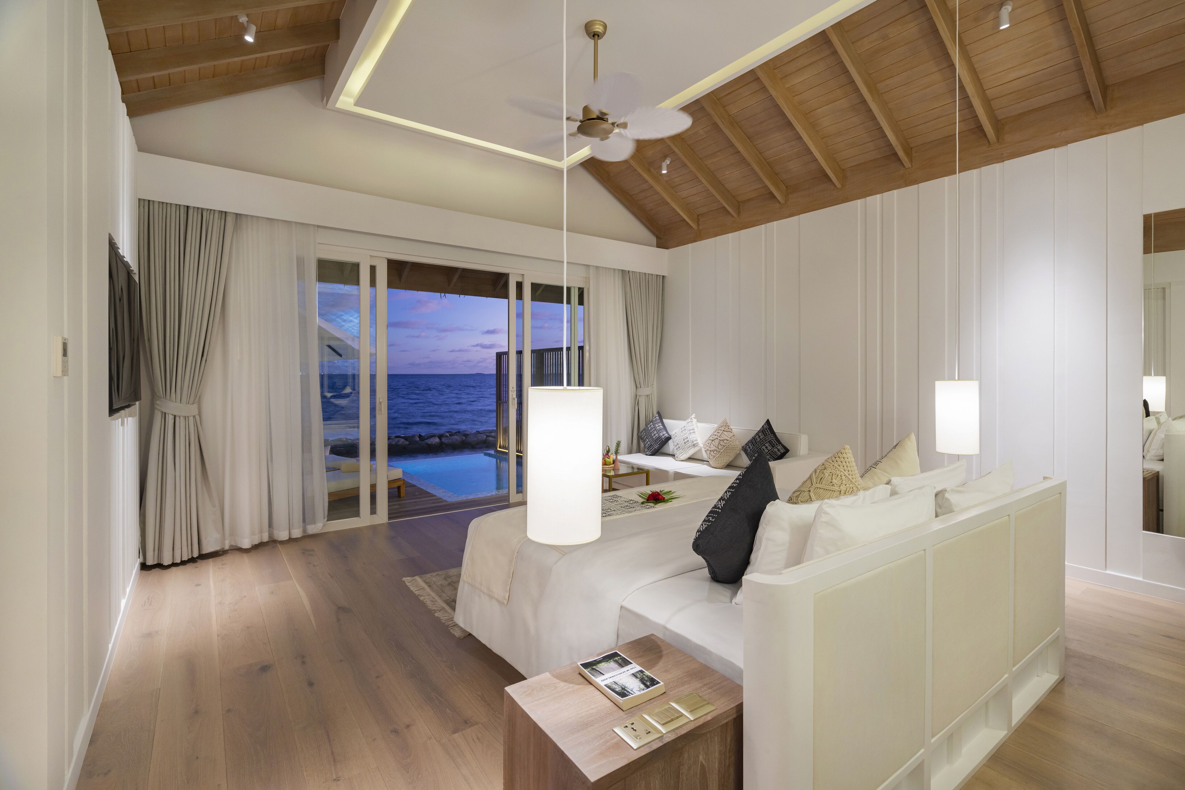 A modern bedroom with a large bed, a ceiling fan, and a view of the ocean through floor-to-ceiling windows.