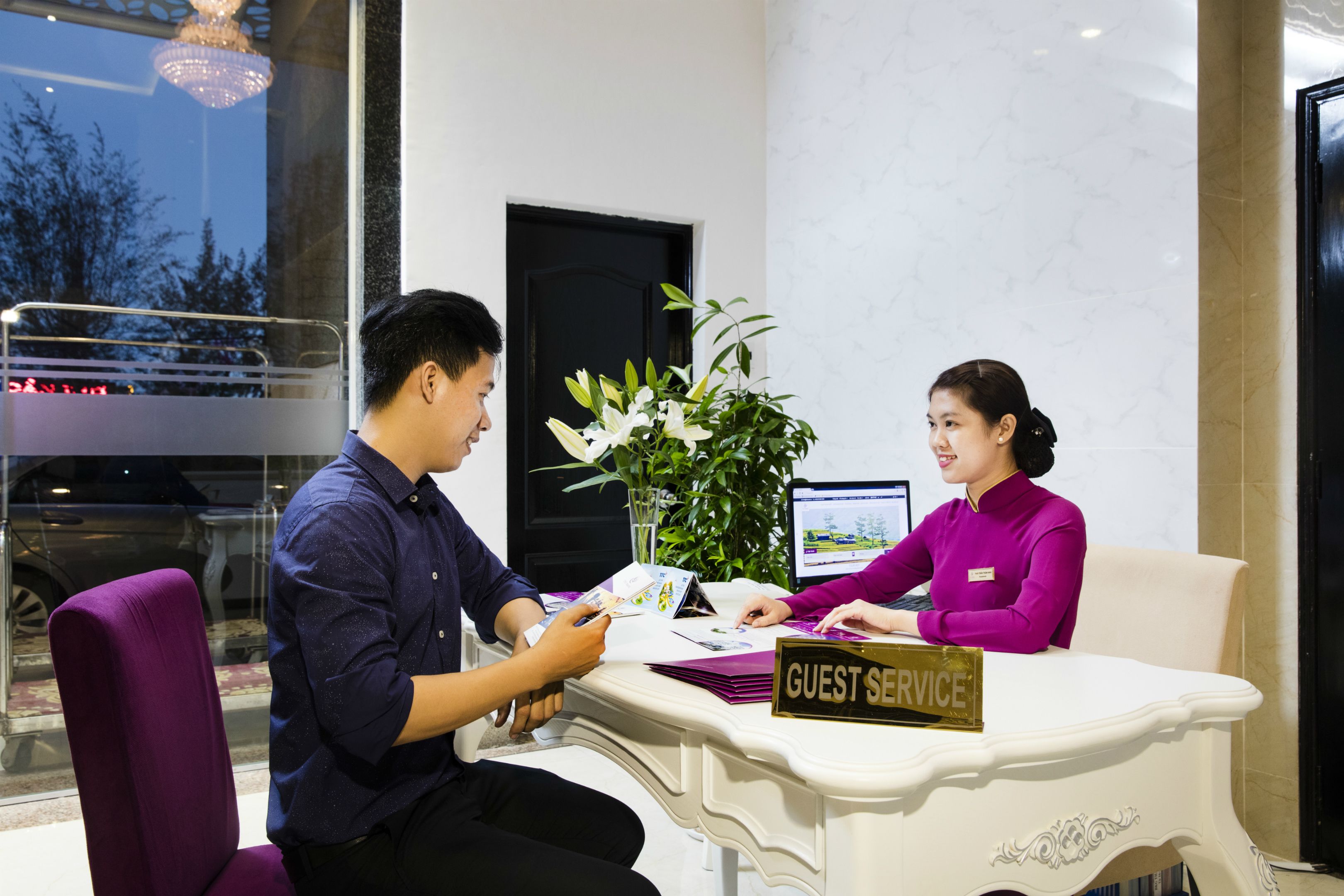 A guest service desk with a sign, a computer, and a plant.