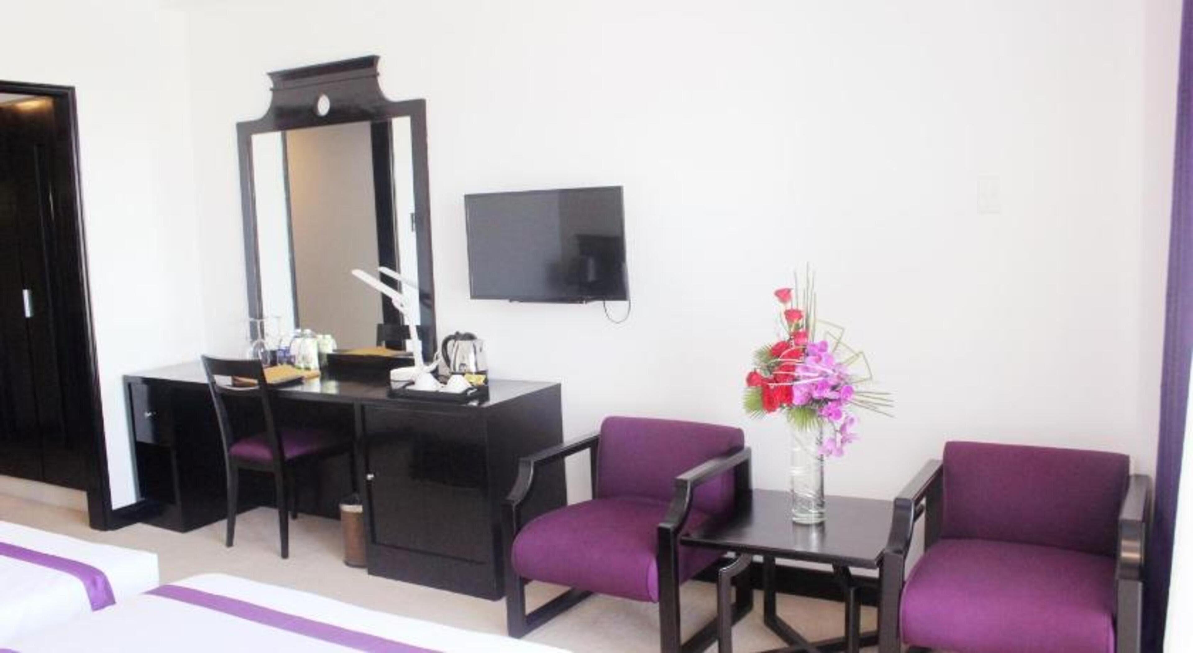 Quarto de hotel com escrivaninha, duas cadeiras roxas, um espelho grande, uma TV de tela plana e um vaso com flores.