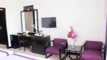 Quarto de hotel com escrivaninha, duas cadeiras roxas, um espelho grande, uma TV de tela plana e um vaso com flores.
