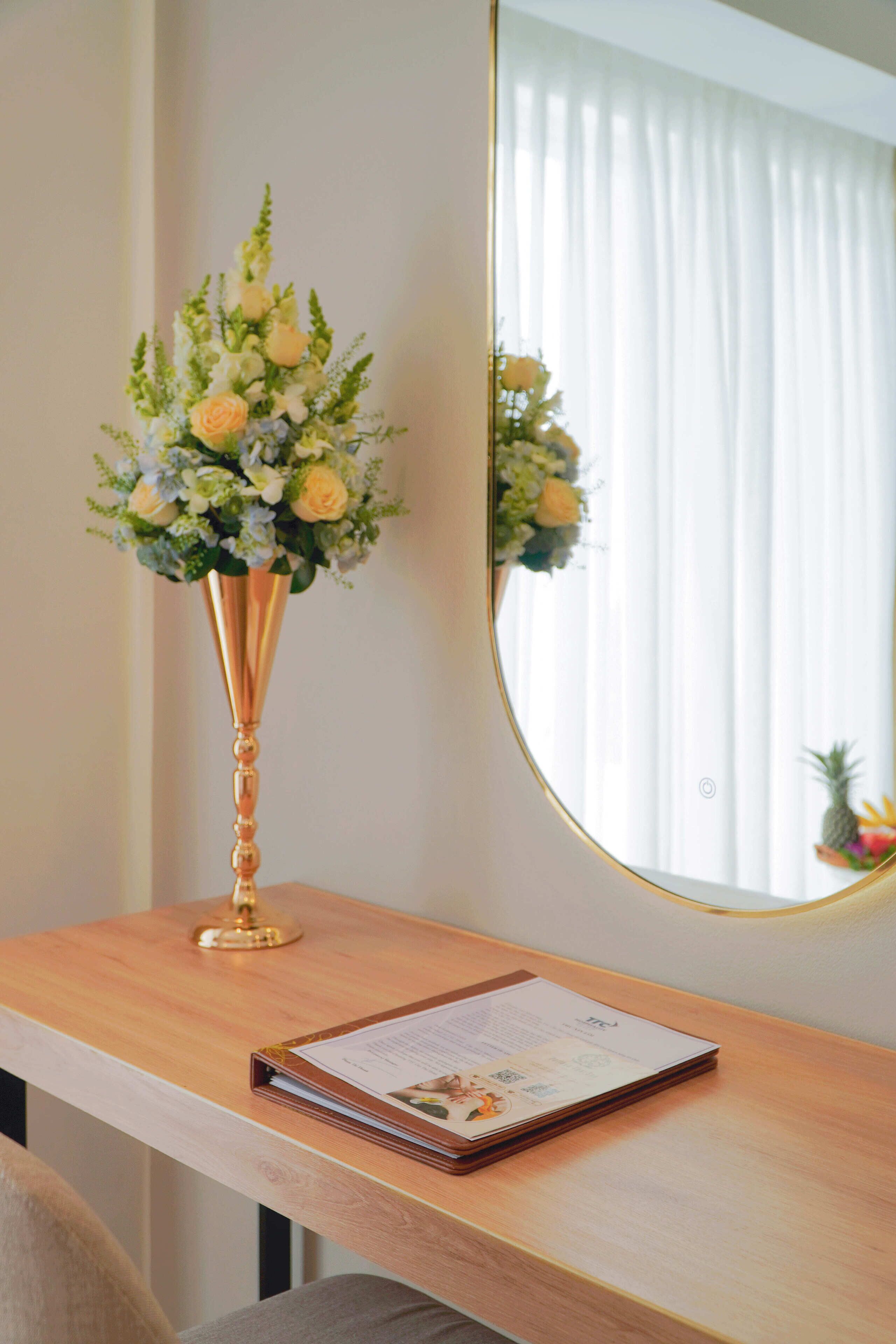Ein Holztisch mit Blumen in einer Vase und ein Spiegel, der ein Fenster mit Vorhängen widerspiegelt.