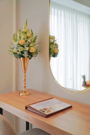 A wooden desk with a vase of flowers and a mirror reflecting a window with curtains.