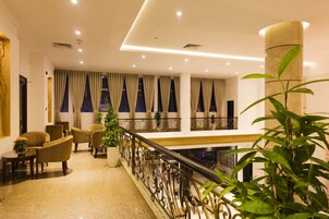 A hotel lobby with a high ceiling, multiple windows with curtains, a balcony with a railing, and several wicker chairs and tables.