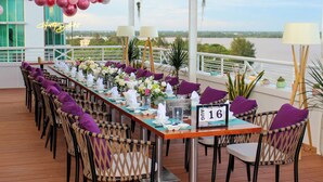 Una larga mesa de comedor con cojines morados, servilletas blancas y centros de mesa florales, rodeada de sillas de mimbre.