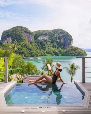 A woman reading by an infinity pool with a view of a large mountain and a body of water.