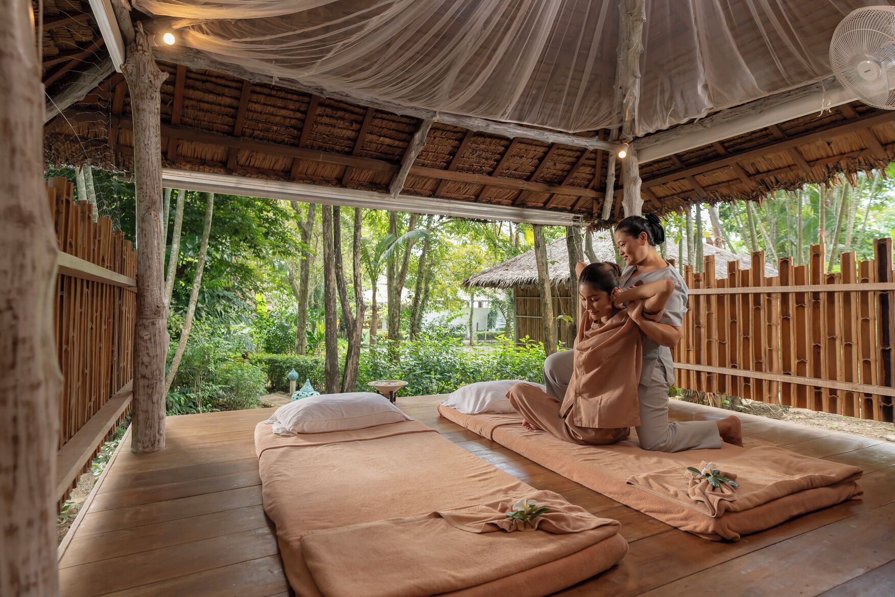 A traditional massage setting with wooden floors and a thatched roof, surrounded by lush greenery.