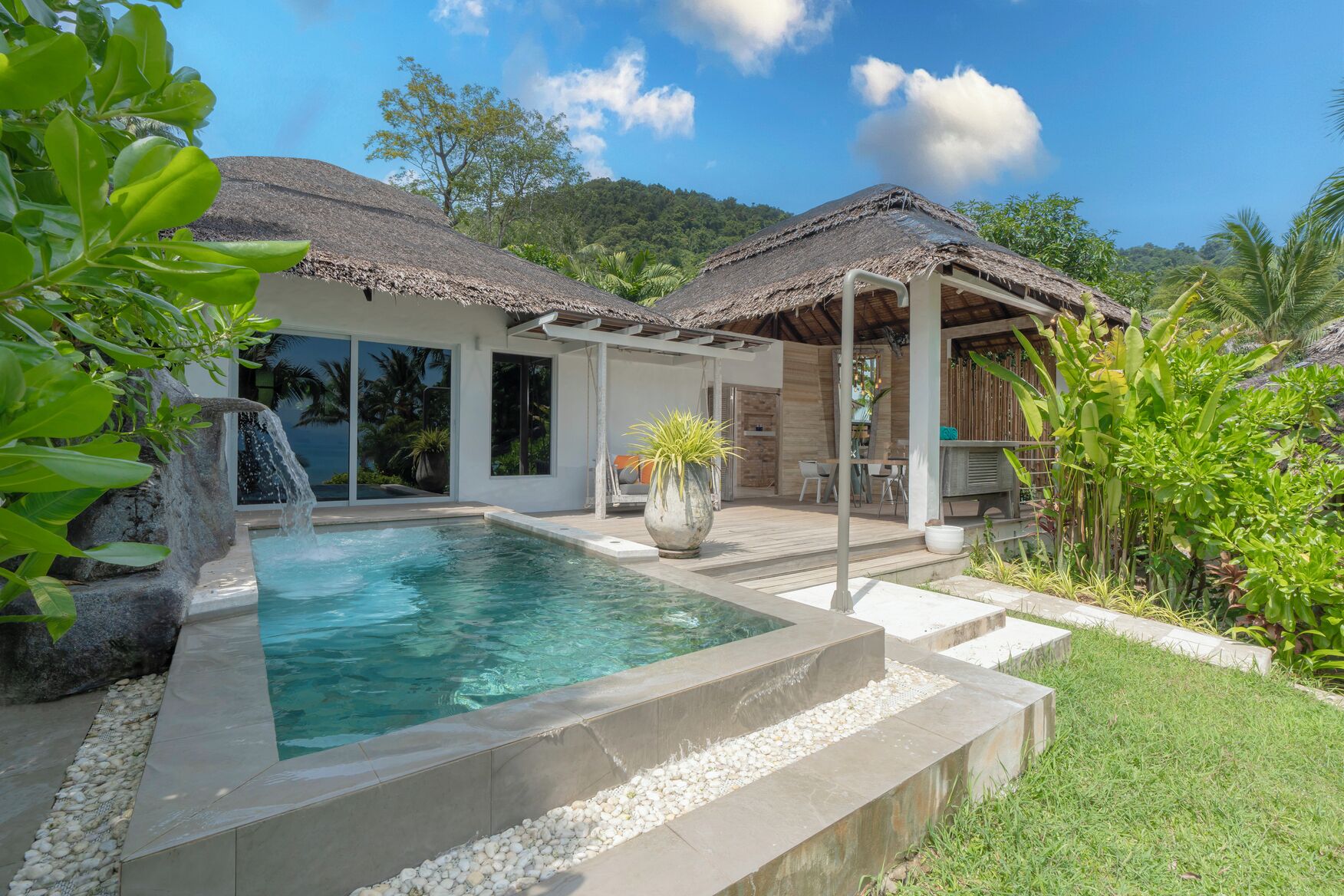 Un área de piscina con una cascada, un espacio de estar cubierto y abundante vegetación.