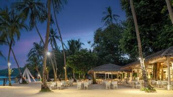 A beachside restaurant with outdoor seating, palm trees, and a view of the ocean.