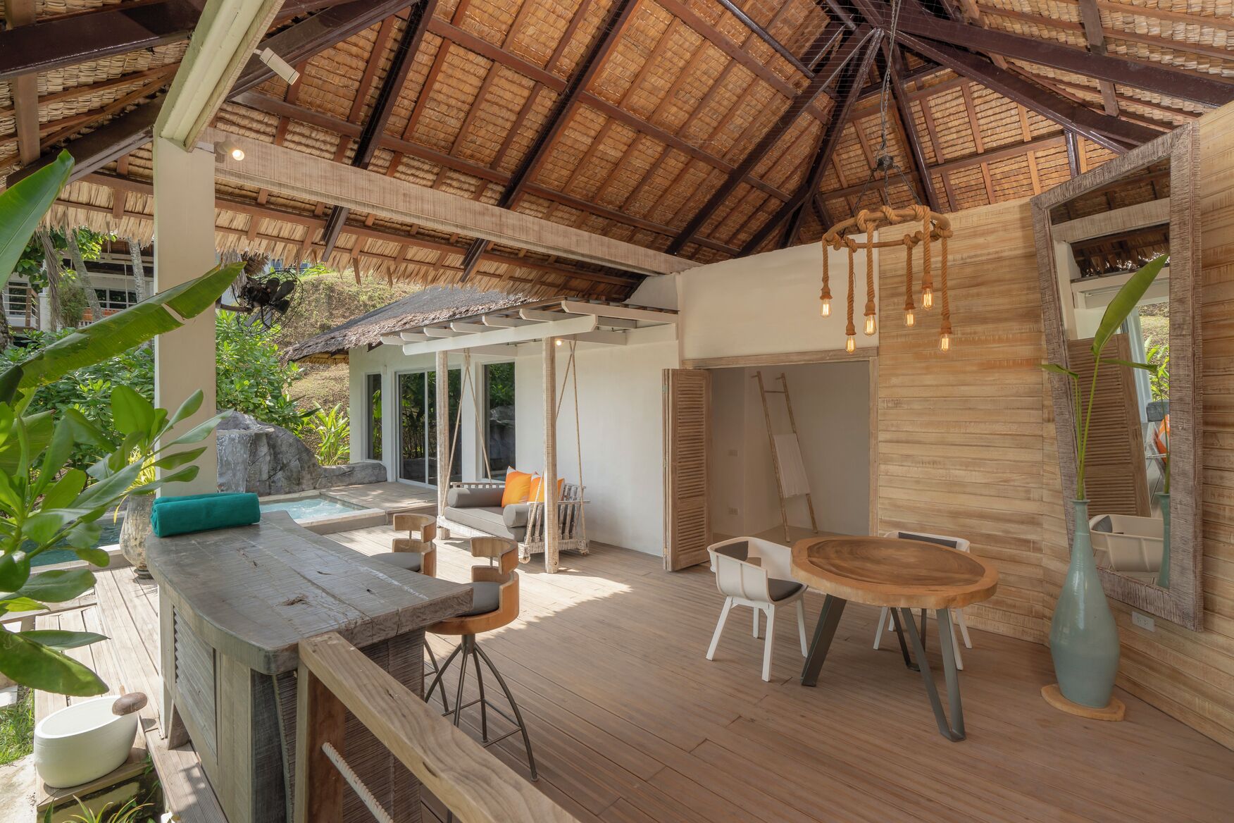 Un espace extérieur aménagé avec une table et des chaises en bois, un luminaire suspendu et une structure dotée d’un toit de chaume.