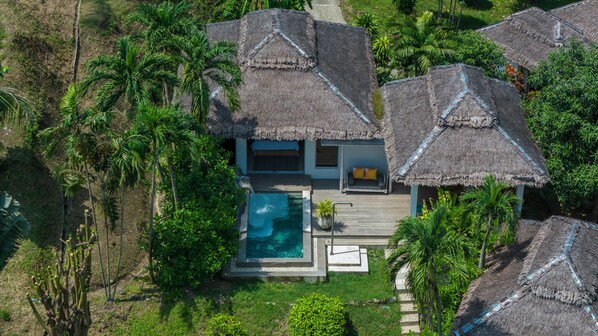 An aerial view of a resort with thatched-roof villas, a central pool, and lush tropical vegetation.