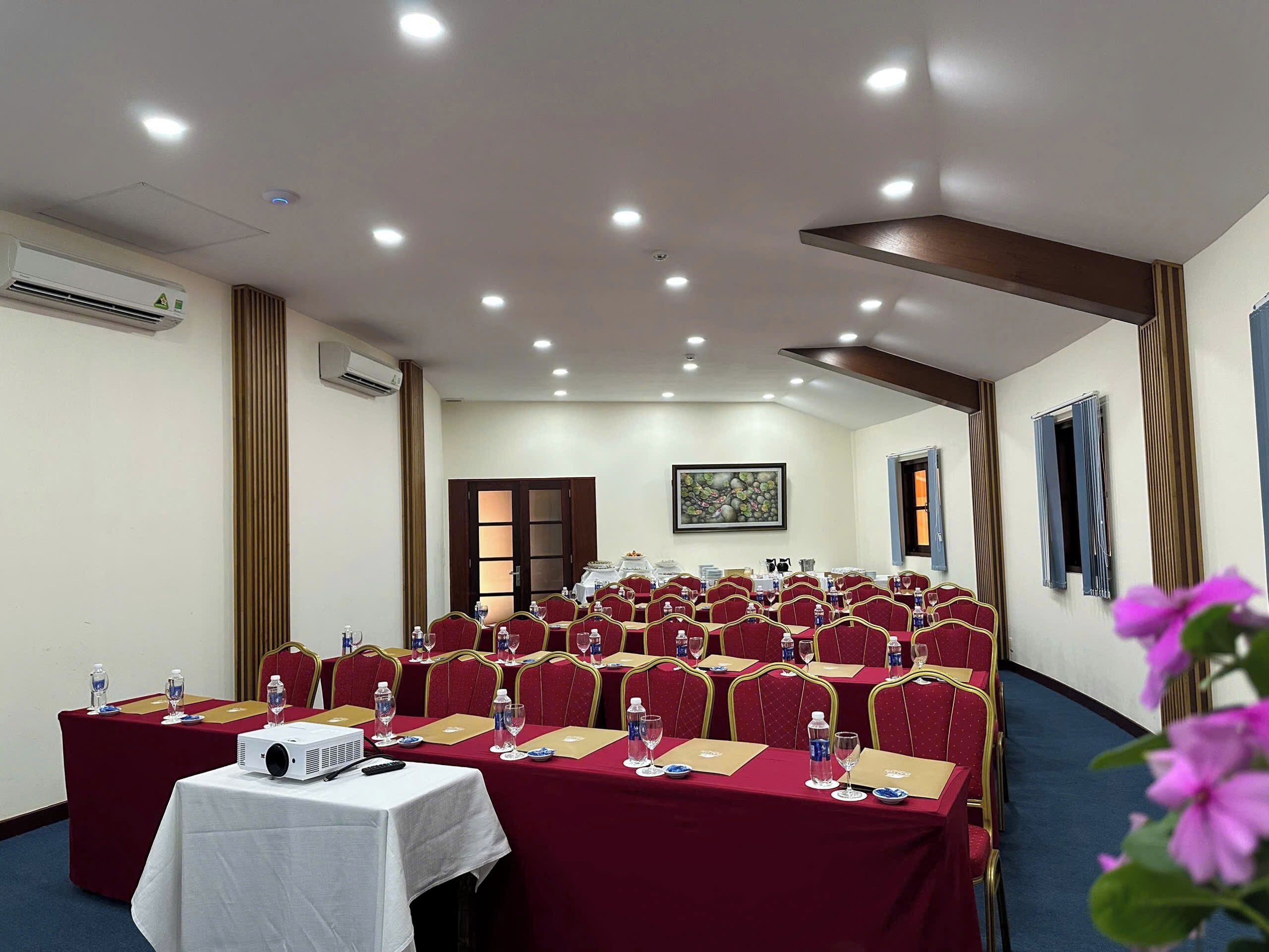 A conference room with rows of red chairs, a white table with a projector, and an air conditioning unit on the wall.