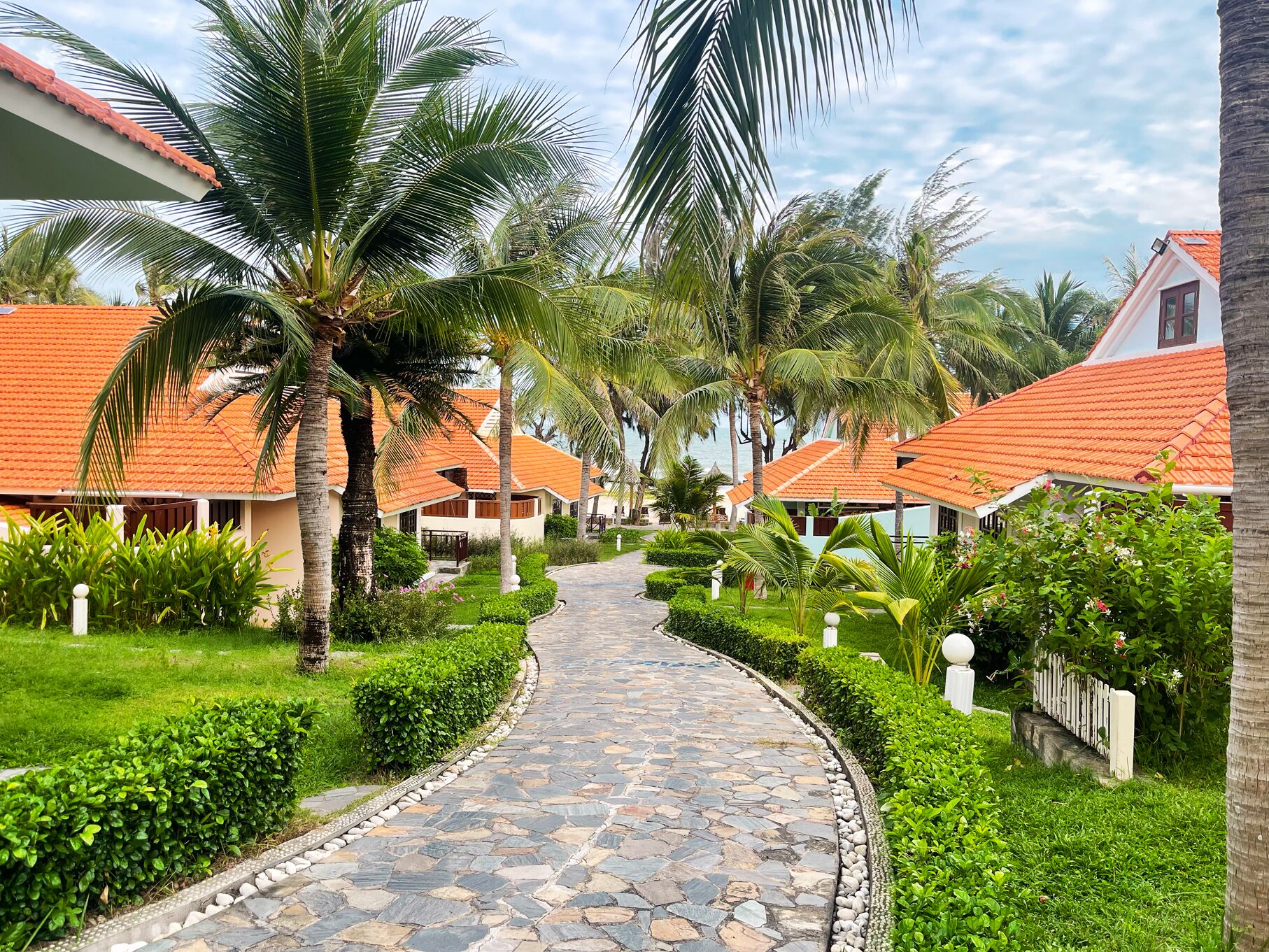 A pathway leading to a row of bungalow-style buildings with red-tiled roofs, surrounded by lush greenery and palm trees.