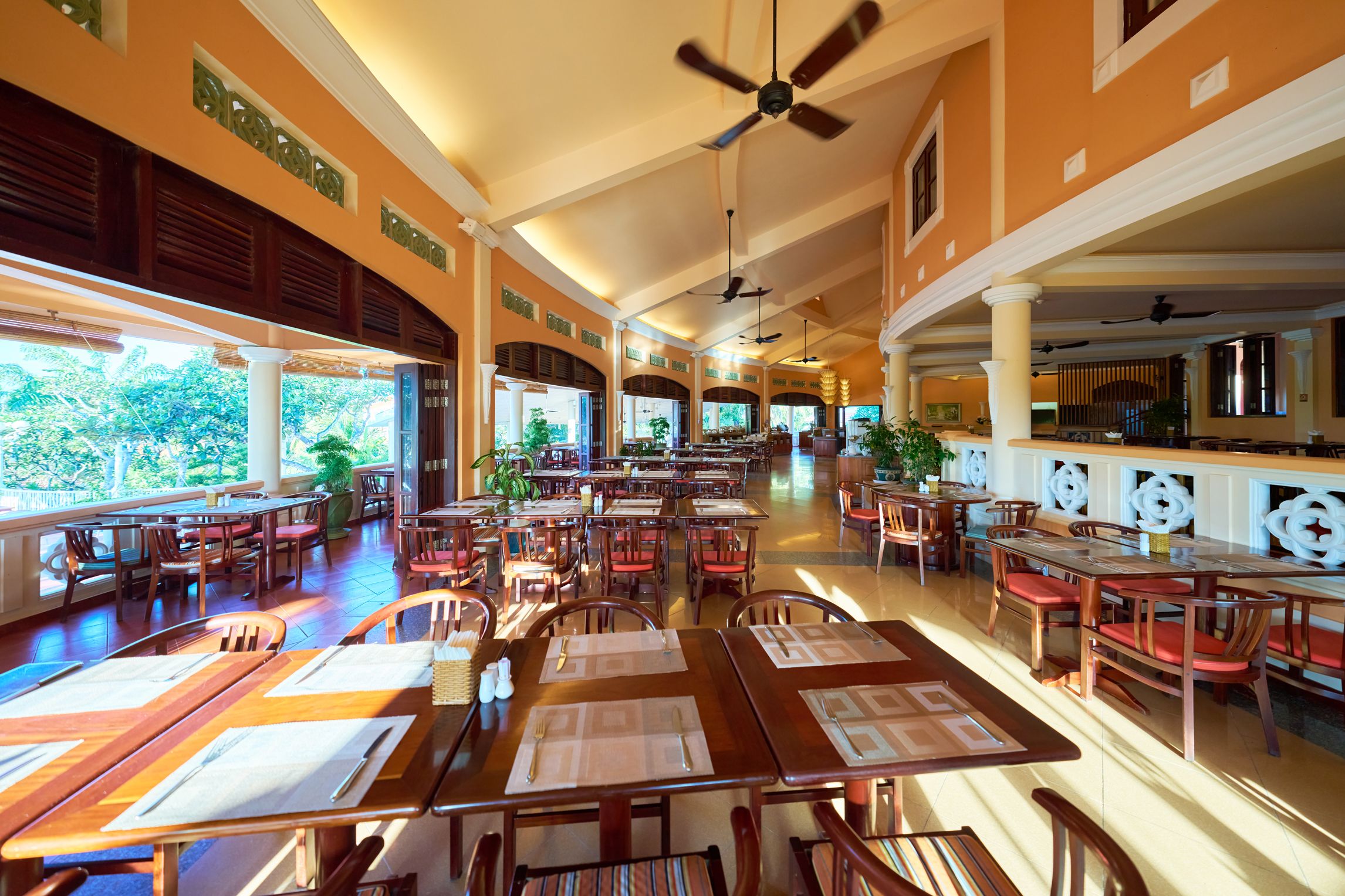 Um restaurante espaçoso com várias mesas e cadeiras, ventiladores de teto e grandes janelas que proporcionam muita luz natural.