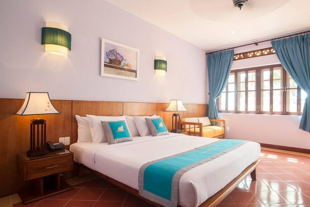 A hotel room with a large bed, two bedside tables with lamps, a wooden headboard, a framed picture on the wall, and a window with curtains.